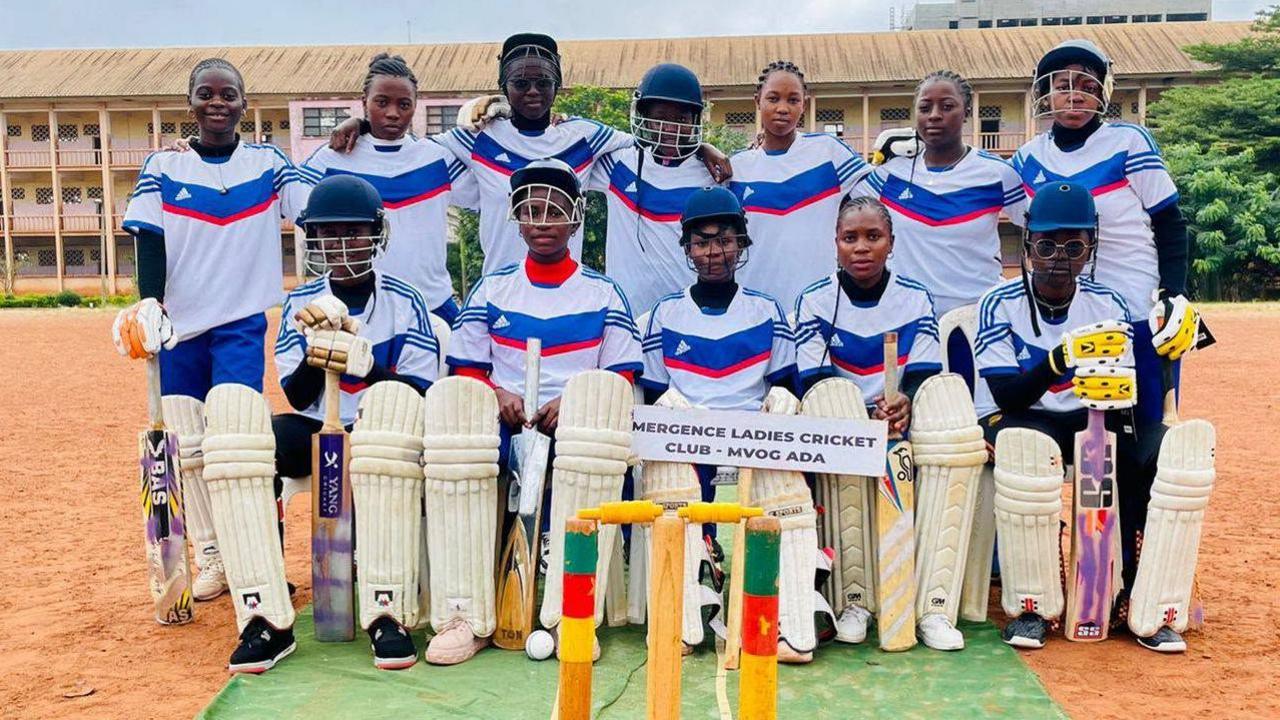 Twelve women line up for a team photo wearing white T-shirts with a broad blue and thin red chevron on the front. Seven are standing in the back row with five players seated in front. The give seated players and the standing player on the far left wear cricket pads on their legs, with seven dotted across the team wearing helmets. The player seated in the middle holds a sign in her lap which reads Emergence Ladies Cricket Club, Mvog Ada. They pose on a green artificial wicket on a clay surface, with stumps bearing the Cameroonian flag colours of green, red and white in front of them. A long three-tiered building can be seen in the far distance
