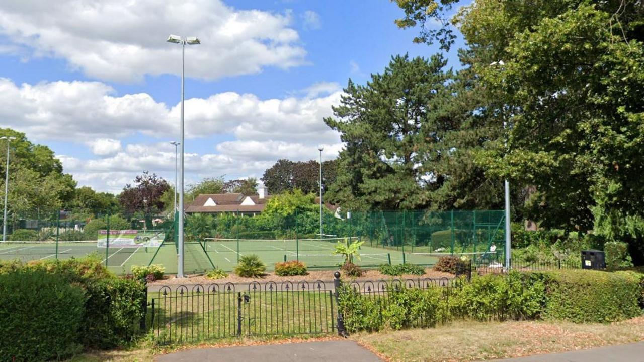 Tennis courts. We are looking from across the road and it is separated by a fence. It is bright day with blue skies and the odd cloud. There are lampposts with lights. We can see trees and some buildings in the background.