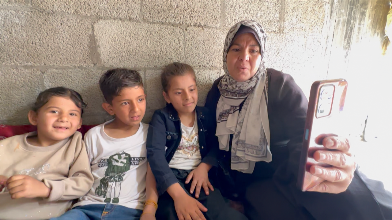 Three children look at a phone next to their grandmother, sitting against a wall