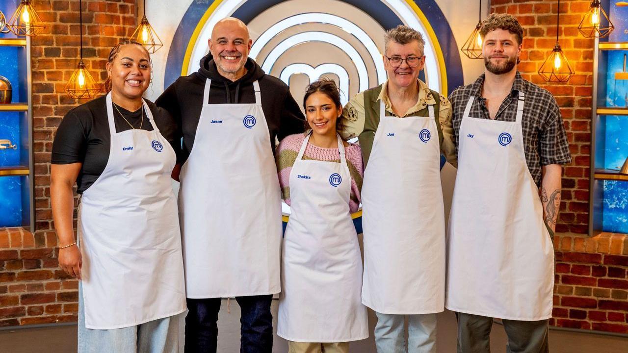 Five people - two women and three men - all wearing white aprons, stand in front of a brick wall.