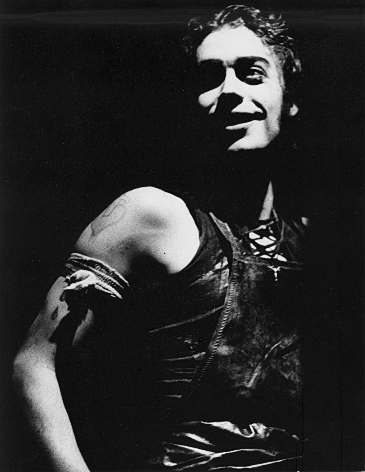 A man dressed in a dark corset on stage. The stage and everything else is entirely dark - only he is illuminated.