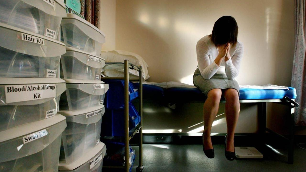 A woman sitting in a private room of a medical building. She is on the right with  her head in her hands, on the left are some storage boxes with medical supplies.