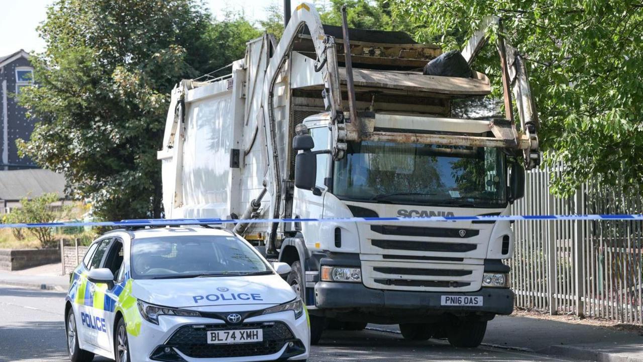 A white police car parked next to a white bin lorry in a street with blue and white police tape in front of them
