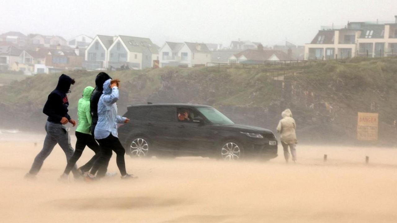 People walking on the beach at Portstewart. Four people in the foreground in blue, black and green coats, with hoods up. There is a black vehicle behind them with a person in a light brown coat to the right. The sand is lifting due to the winds. There are buildings in the distance.