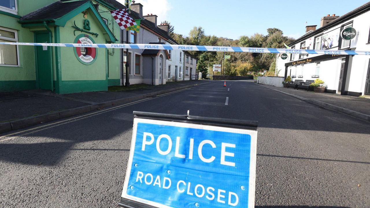 A police road closed sign with white writing on a blue background sitting in the middle of the road.
There is police tape tape across the street above it.
On one side of the street there is a green building and on the other side a white pub with a Carlsberg sign sticking out.