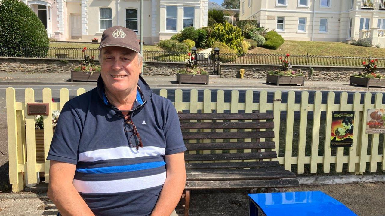 A man in a blue top with white and blue stripes. He is wearing a brown cap. Behind him is a bench. There is a road with flowerpots on the pavement. There are white houses in the background.