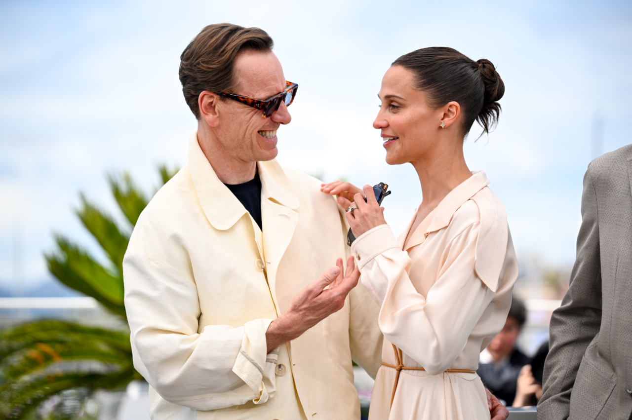Two people, a man and a woman, stand together outdoors. The man on the left wears a light coat and dark sunglasses, angled slightly toward the woman as if mid‑conversation. His posture is open, comfortable, and engaged. The woman on the right is dressed in a soft beige outfit, holding a pair of sunglasses loosely in one hand. Her expression looks warm and attentive, suggesting an easy, friendly exchange. Behind them is bright greenery and a clear sky.