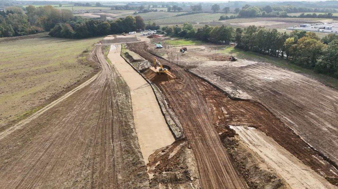 Drone image of the earthworks, with a digger in the middle, and fields and trees bordering the site.