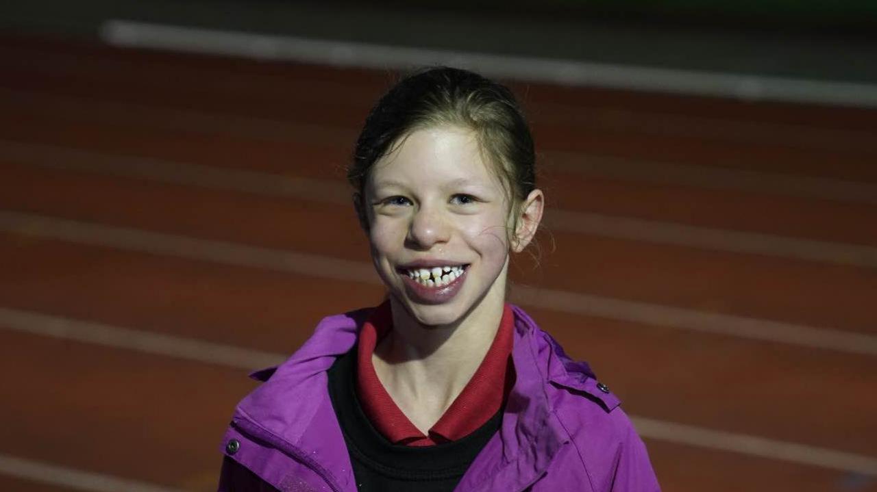 Fabrienne smiles widely at the camera while standing on the athletics track. She is wearing a purple coat with a black jumper and red polo top on underneath. She has long brown hair which has been tied up behind her head.