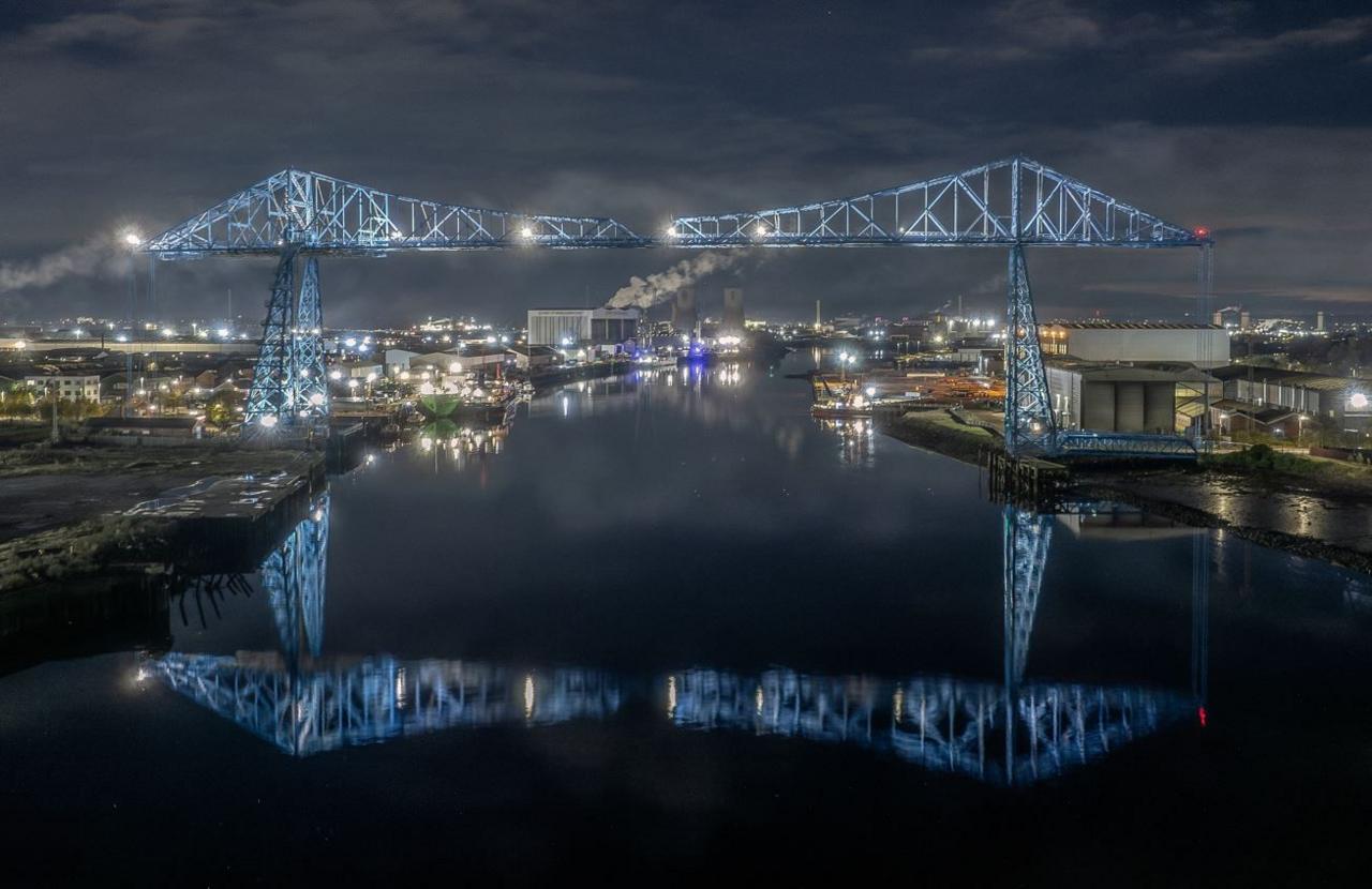 A metal blue bridge at night reflected in the dark river below it. City lights can be seen stretching into the distance, with smoke from industrial chimneys.