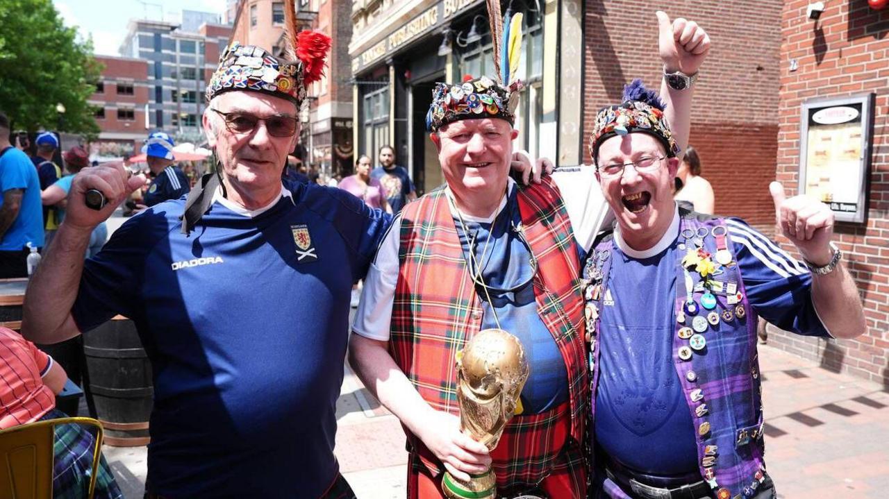 Three Scotland fans in Scotland shirts, one is holding a replica World Cup trophy