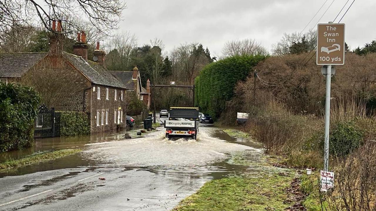 A tipper van driving away through flooding in a country lane. to the left is an old brown brick building