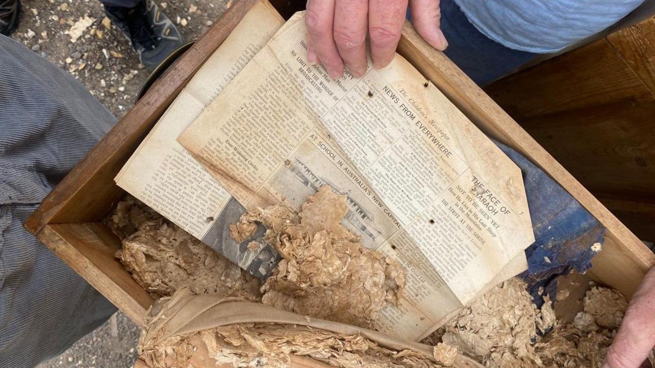 A close up of the stool after the cover was removed. Inside the wooden frame is some old wadding and a well-preserved copy of the Children's Newspaper. One of the headlines reads: The Face of a Pharaoh.