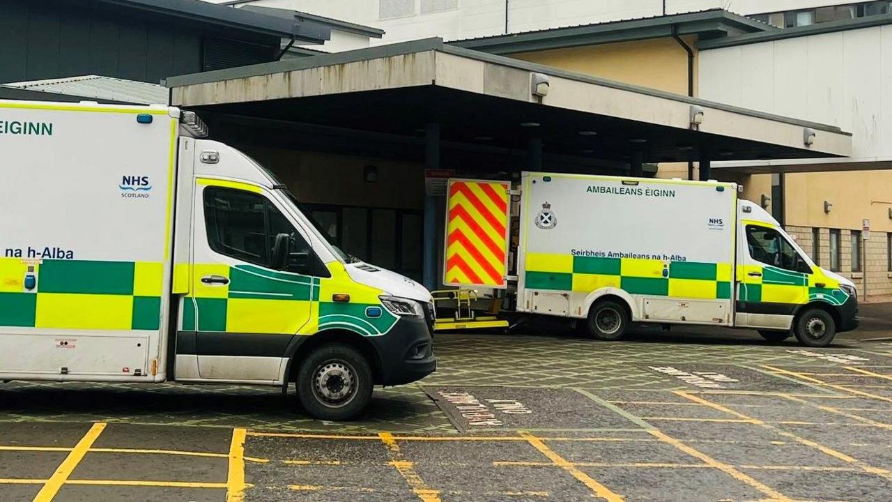 Ambulances with green and yellow checked markings, parked outside a hospital building.