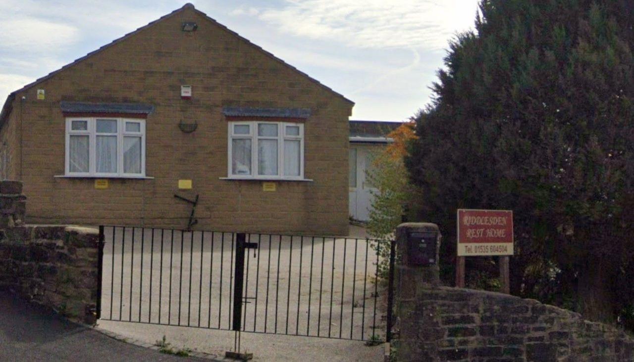 A screenshot of a former care home. In front of the home is a closed black gate and a paved driveway, alongside a sign bearing the home's name.