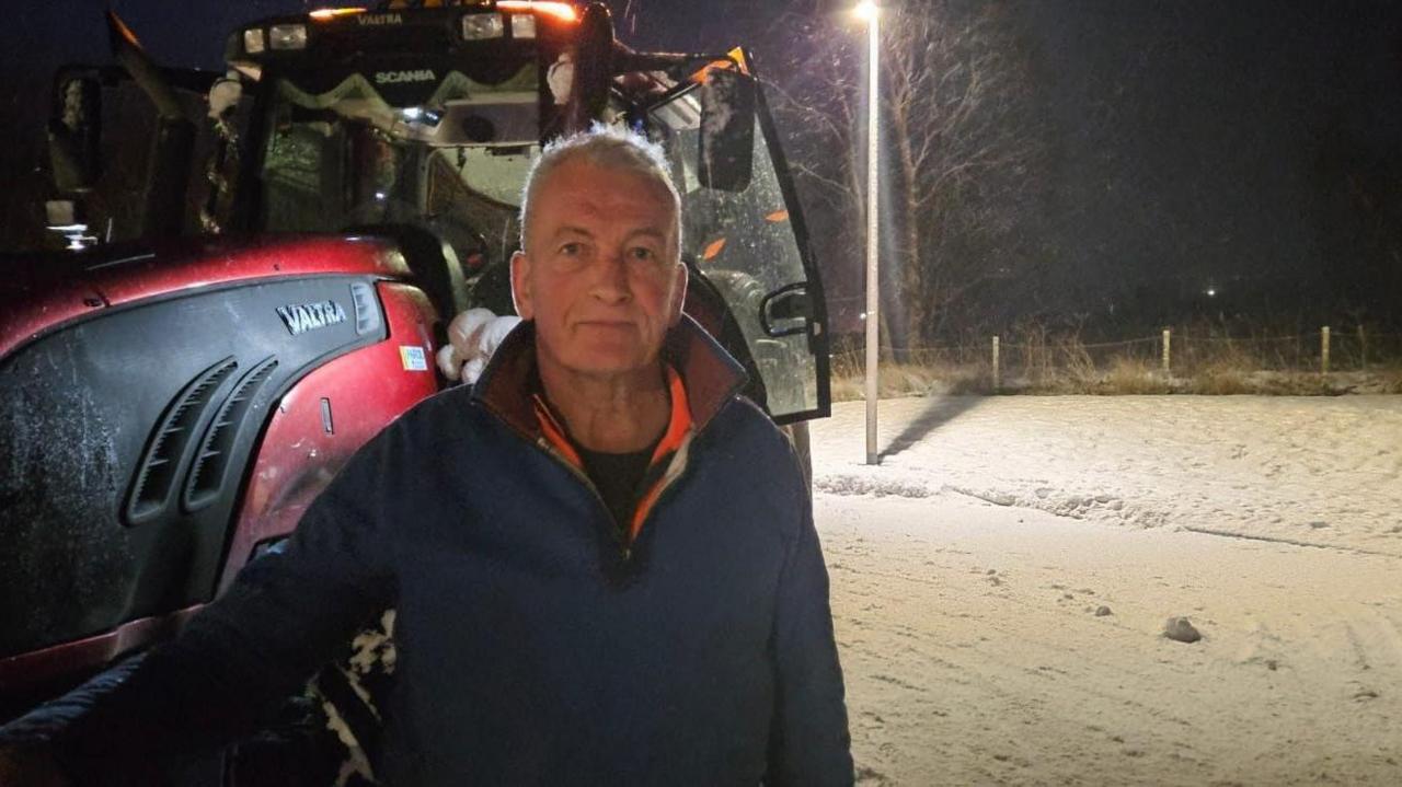 A man with white hair wearing a blue fleece stands in front of a snow plow and a snowy road.