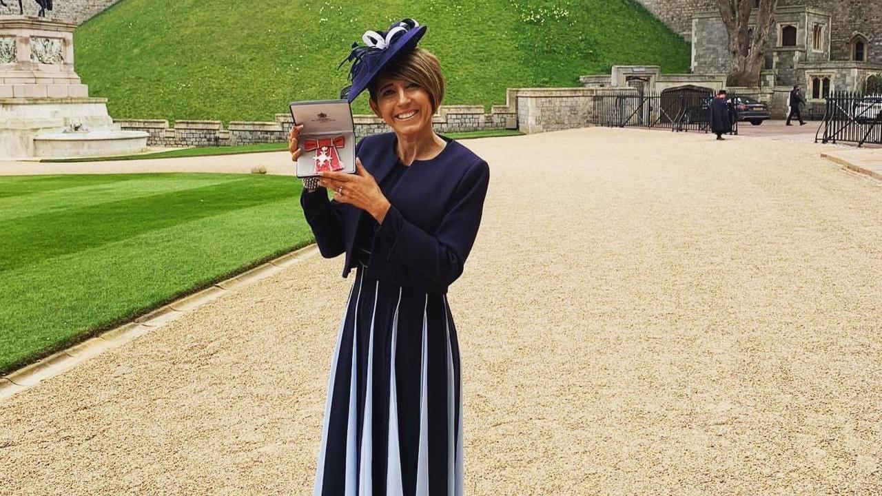 A woman with short brown hair is wearing a navy blue dress with white slits. She is wearing a navy and white hat. She is holding a red MBE medal in a box, and is standing in front of a green garden