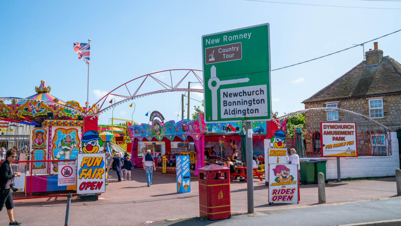 Dymchurch Amusement Park on a sunny summer day, advertising fairground rides. In the foreground is a road sign with directions to New Romney, Newchurch, Bonnington and Aldington.