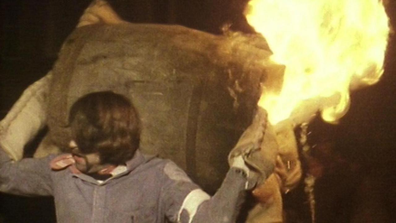 A barrel roller in 1986 hoists a barrel onto his back, he is wearing a long sleeve blue jacket and big sacking gloves. The barrel behind him is on fire.