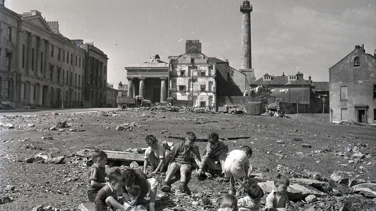 A black and white photo shows children playing on a former demolition site in Devonport. The houses have been cleared after bombs destroyed most of the street.