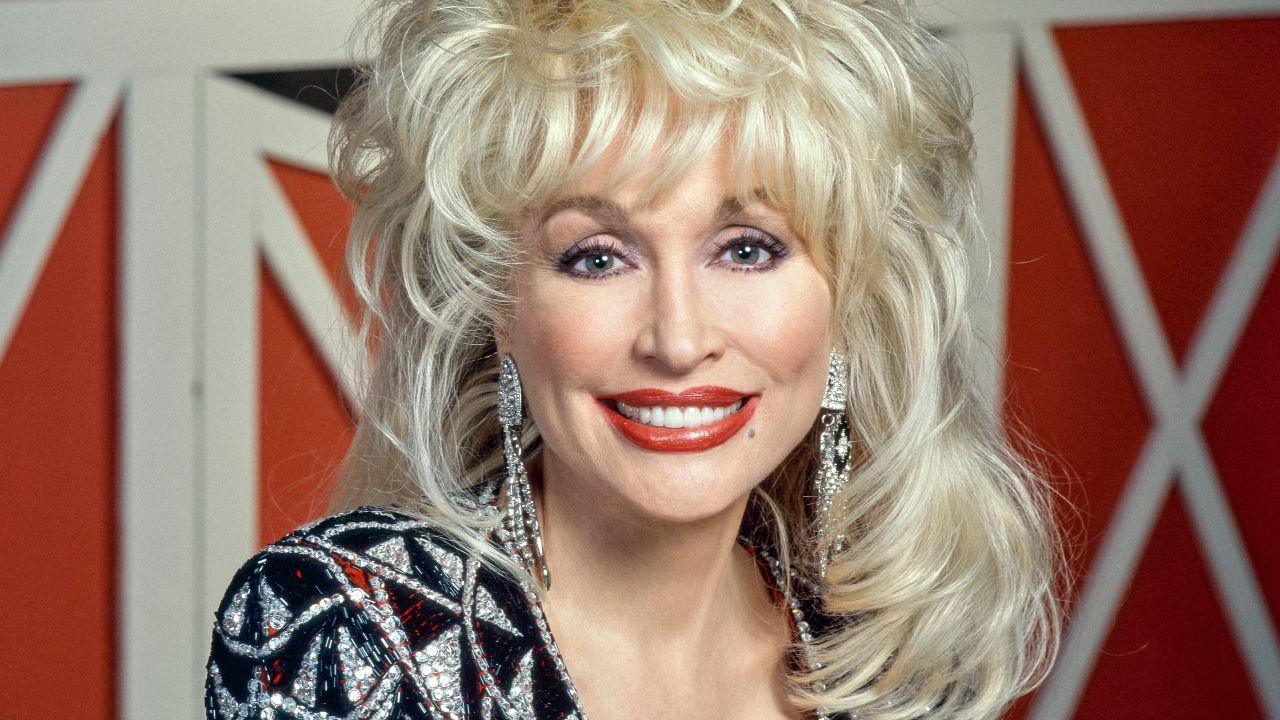 A head and shoulders shot.  Dolly's blonde hair stands up in big waves and falls onto her shoulders.  She wears a sliver and black diamante V neck dress with red accent colours.  Long diamante earrings hang down.