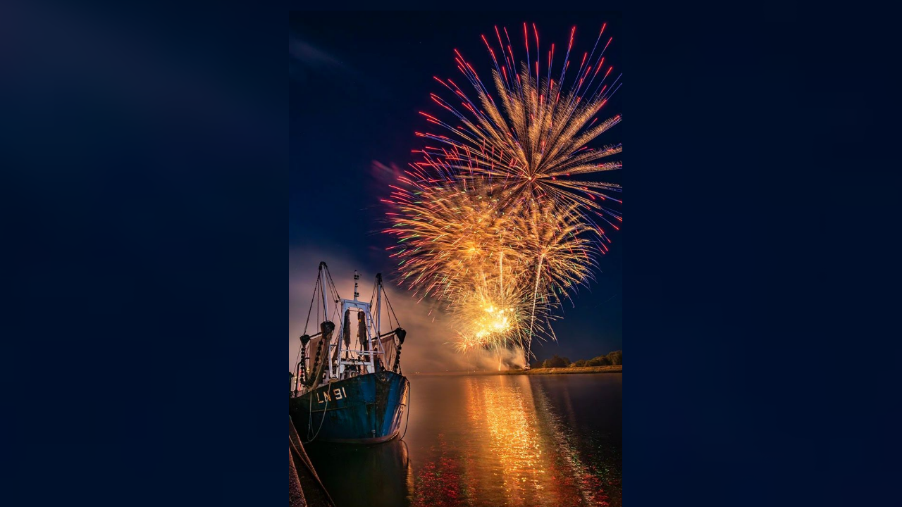 A fishing boat is in the foreground backlit by fireworks being launched from the bank