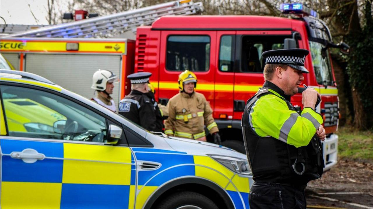 A police officer in his uniform speaking into a radio in the foreground. In the background there is a police car and fire engine, with two firefighters speaking to a policeman. 