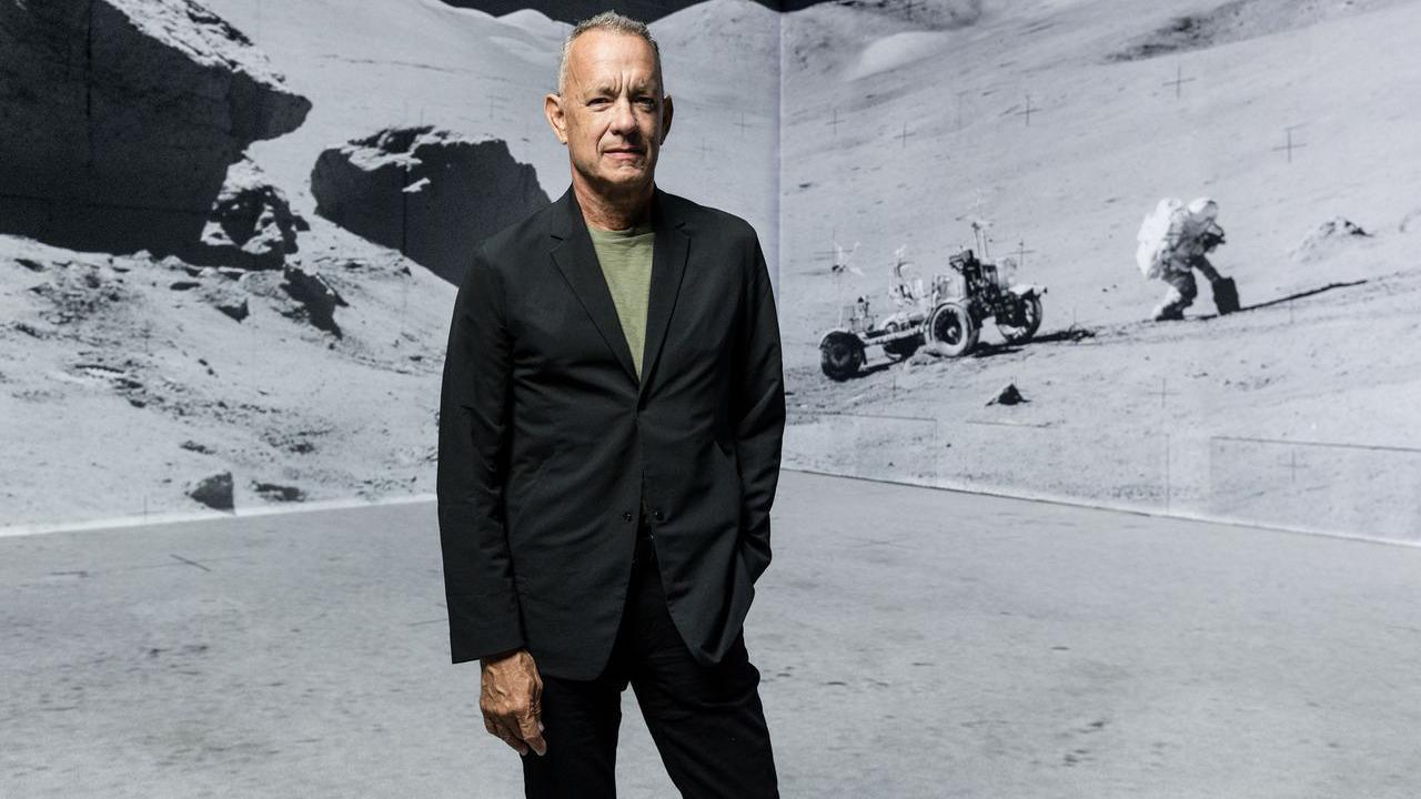 Tom Hanks in black suit and green T-shirt stands in front of digitally remastered shot of an astronaut and the Moon Rover on the surface of the moon.