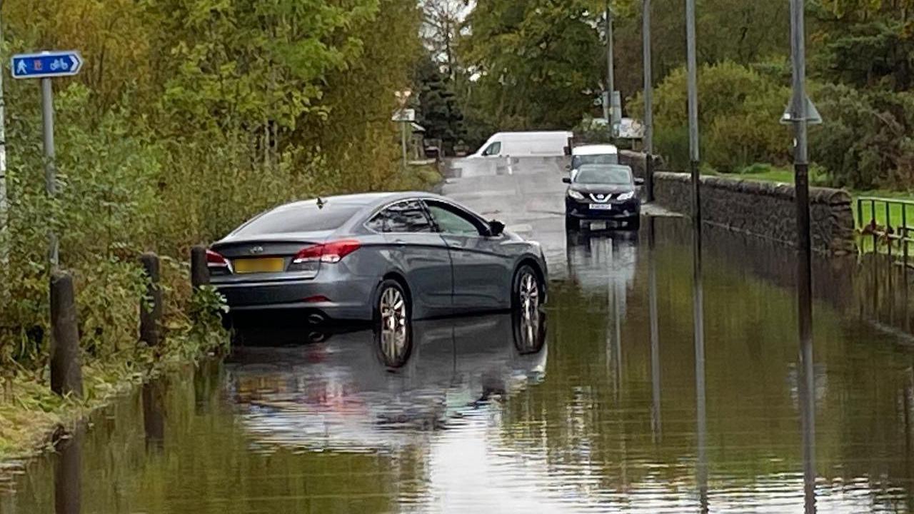 A flooded road with a grey car which appears to be stuck and is at an angle across the road.