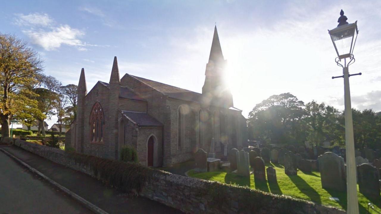 St Peters Church in Onchan, which is a rectangular sandstone church building with a spire at the far end. It is surrounded by a churchyard covered with green grass and headstones. There is also a Victorian-style lamppost in the fore.