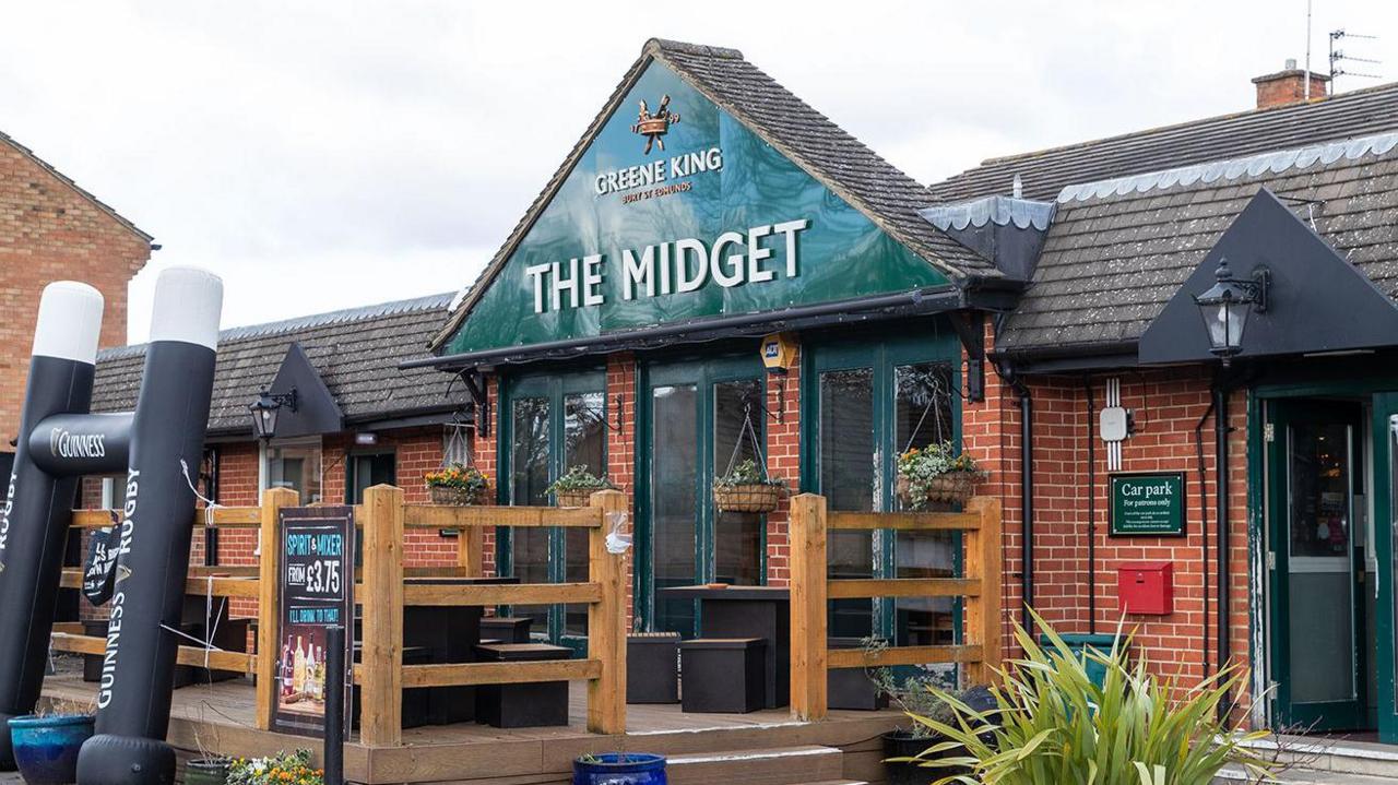 The front of The Midget pub in Abingdon. It is red brick with an arch like entrance. The pub's name is in large white letters on a green background