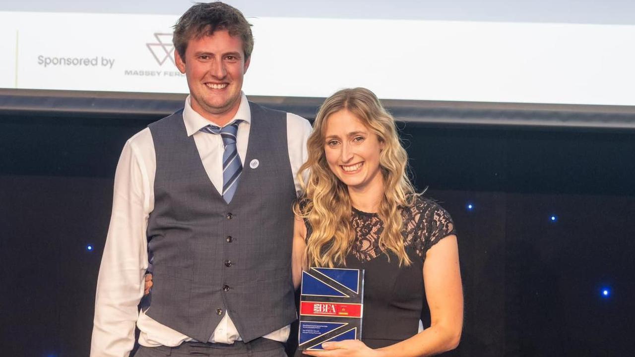 Sam and Samantha Edwards posing with their award. Sam has short blond hair and is wearing a grey waistcoat and trousers with a white shirt and blue tie. Samantha has long blonde hair and is wearing a black dress with lace on the top of the bodice and short sleeves.