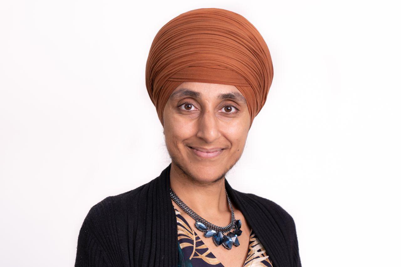 A woman wearing an orange turban smiles at the camera. She has a large blue necklace and a black cardigan on over a dress. The woman stands in front of a white background and the photo is a close-up of her head and shoulders.