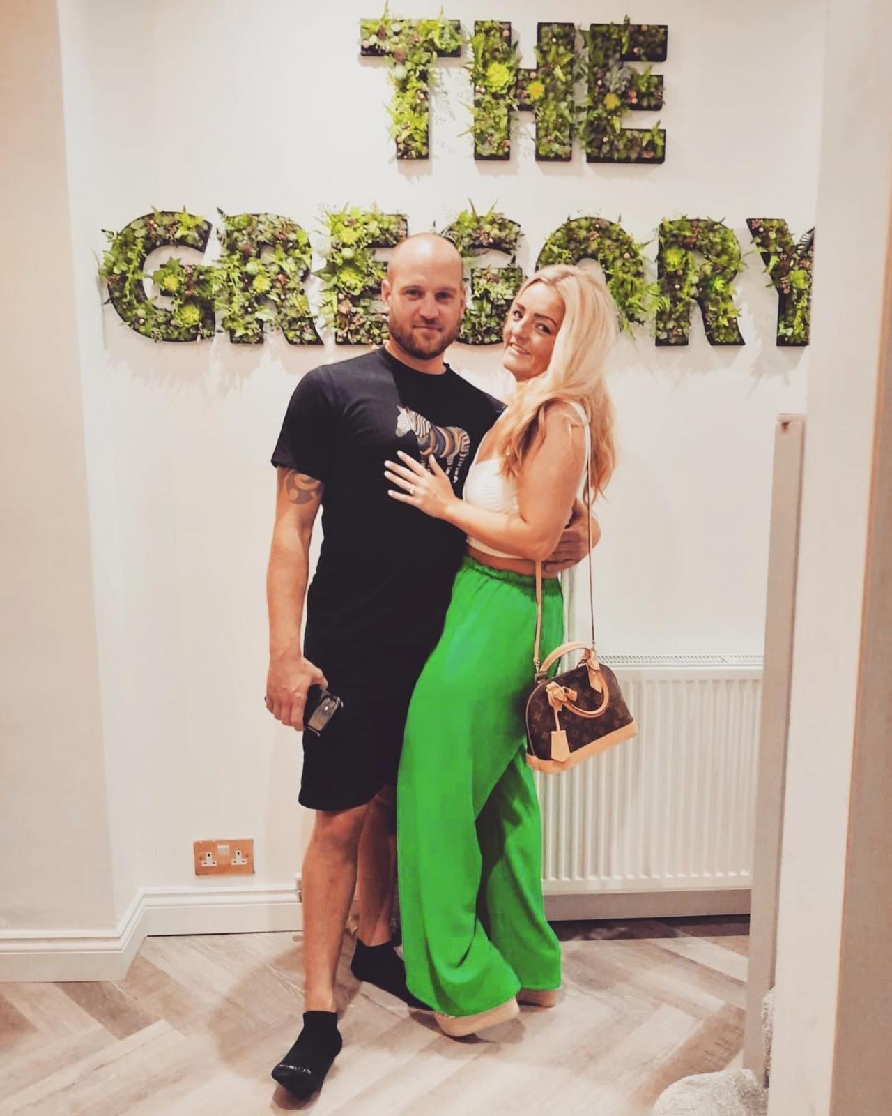 Couple looking happy and smiling for the camera, with a sign behind them which reads "The Gregory". She is wearing pea-green trousers and a white vest top and he is in black shorts and T-shirt