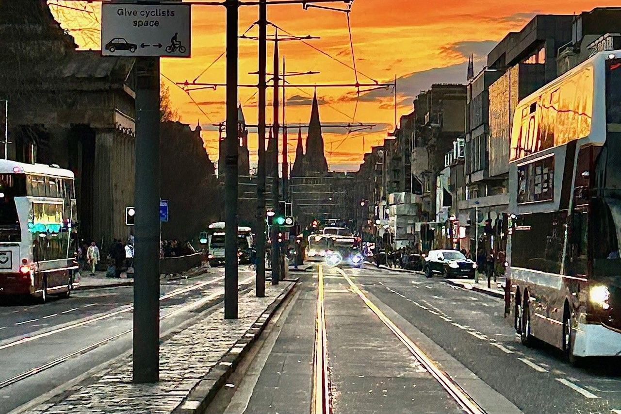 The sky is orange as the sunset falls over the street. There are buses and cars.