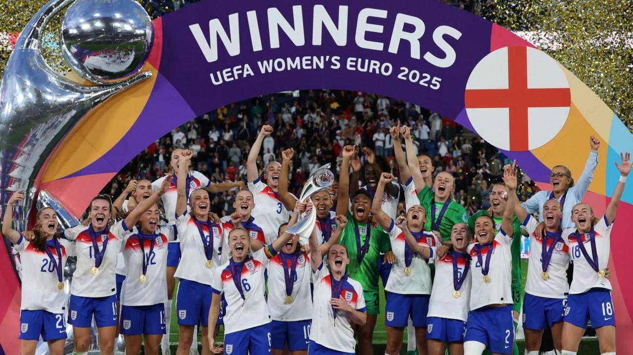 The Lionesses lift the trophy at the European Championship in Basel, confetti is flying behind the players and the team cheers.