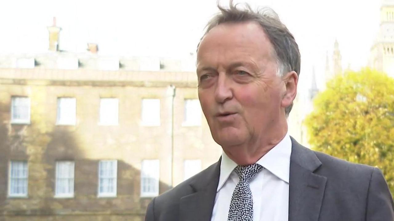 The image shows Andrew George in a dark suit and black and white spotted tie standing outdoors, with a backdrop that includes trees and a historic building with rows of white sash windows.