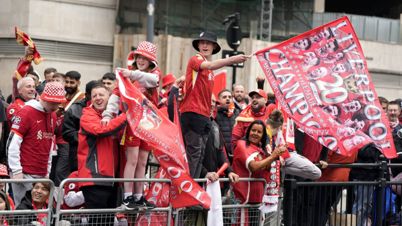 Liverpool fans lining the streets. There are children stood on railings waving flags. With many of the fans in red t-shirts.