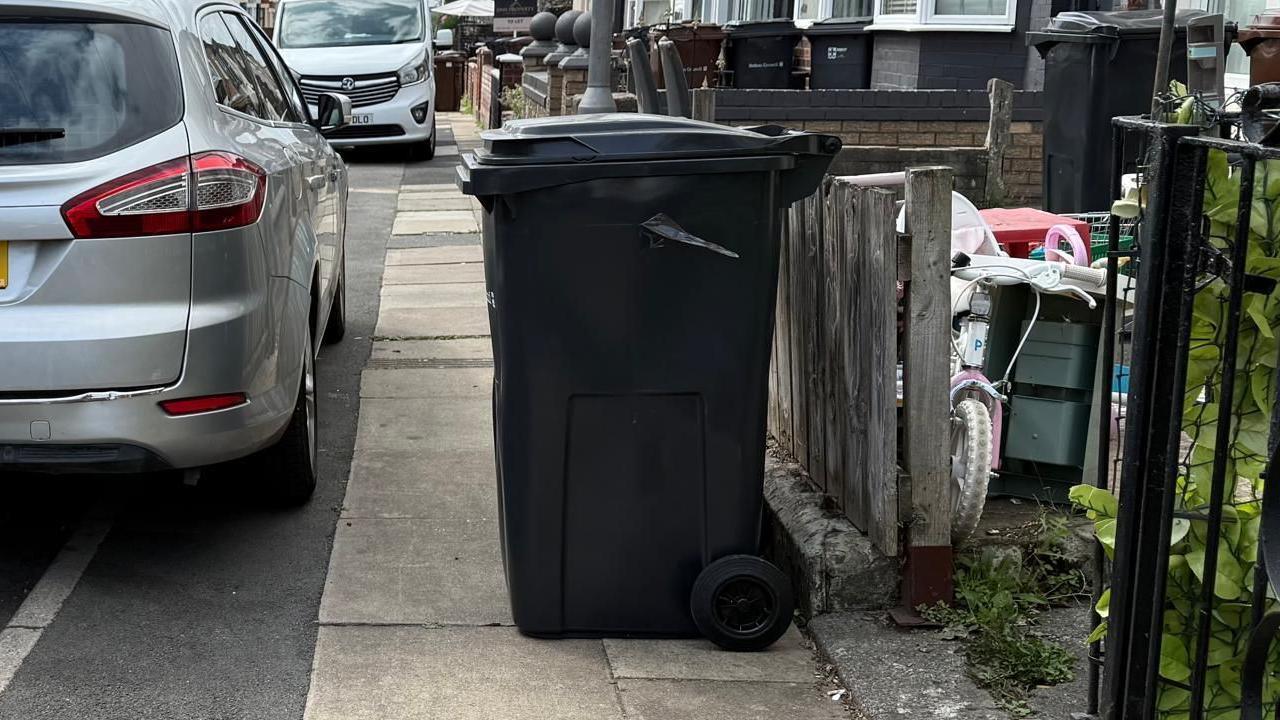 A black wheelie bin is on the pavement on the street. A car is partially parked on the pavement next to it.