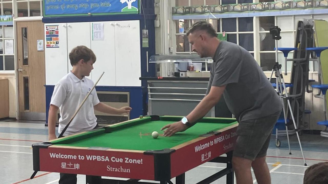 A child in a white polo top holding a cue receives instruction from Kyren Wilson as they stand over a small snooker table.
