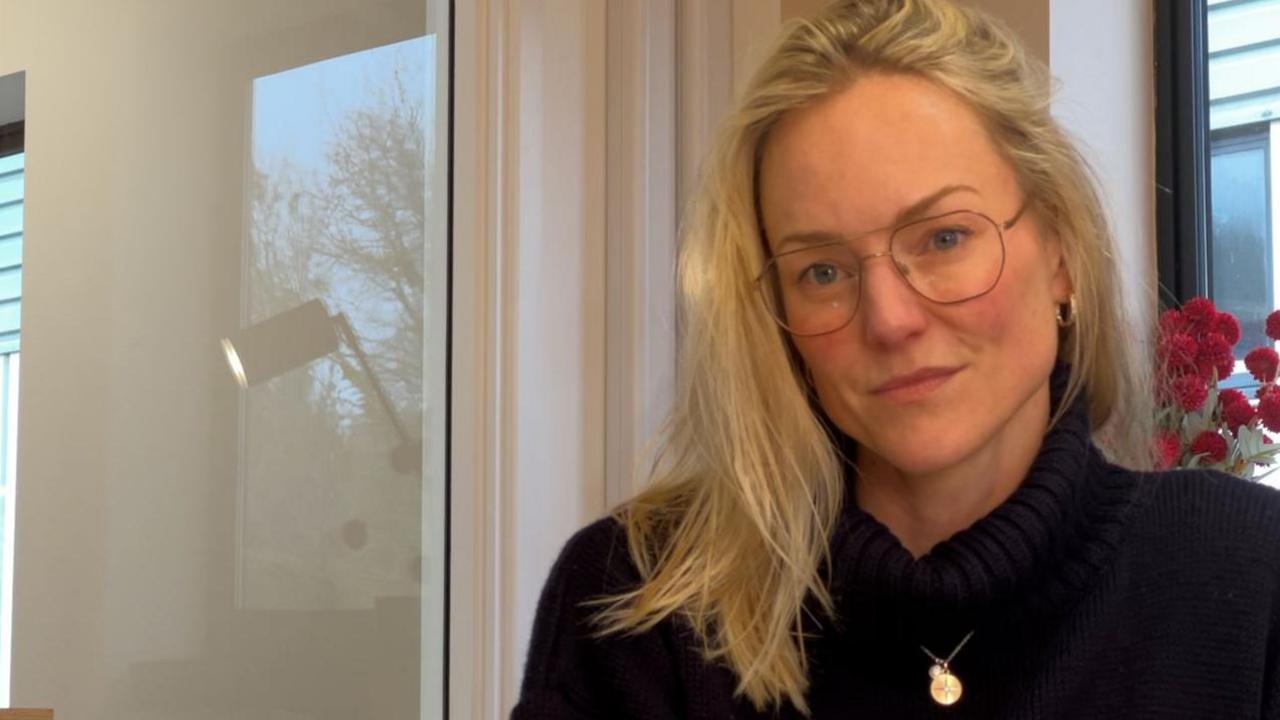 A woman with blonde hair and glasses who is wearing a black jumper and is looking into the camera.