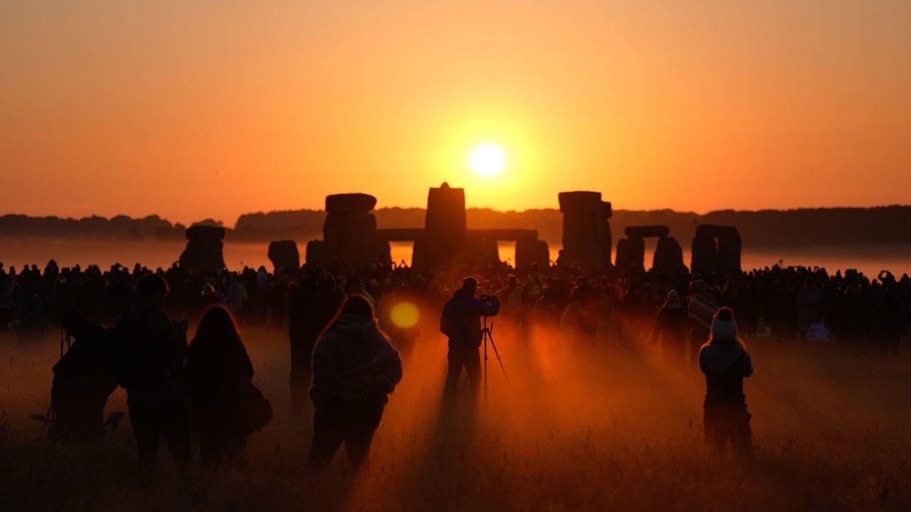 People take pictures and watch the sunrise above the stones at Stonehenge, with the light from the Sun filling the sky orange