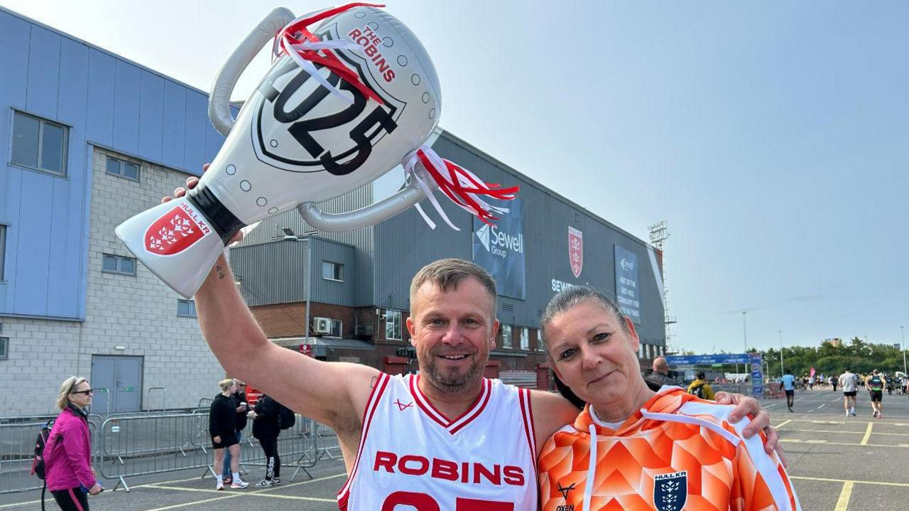 Chris wears a Robins white and red vest and holds up a giant blow up trophy with the Robins 2025 on it. His arm is around his female friend.