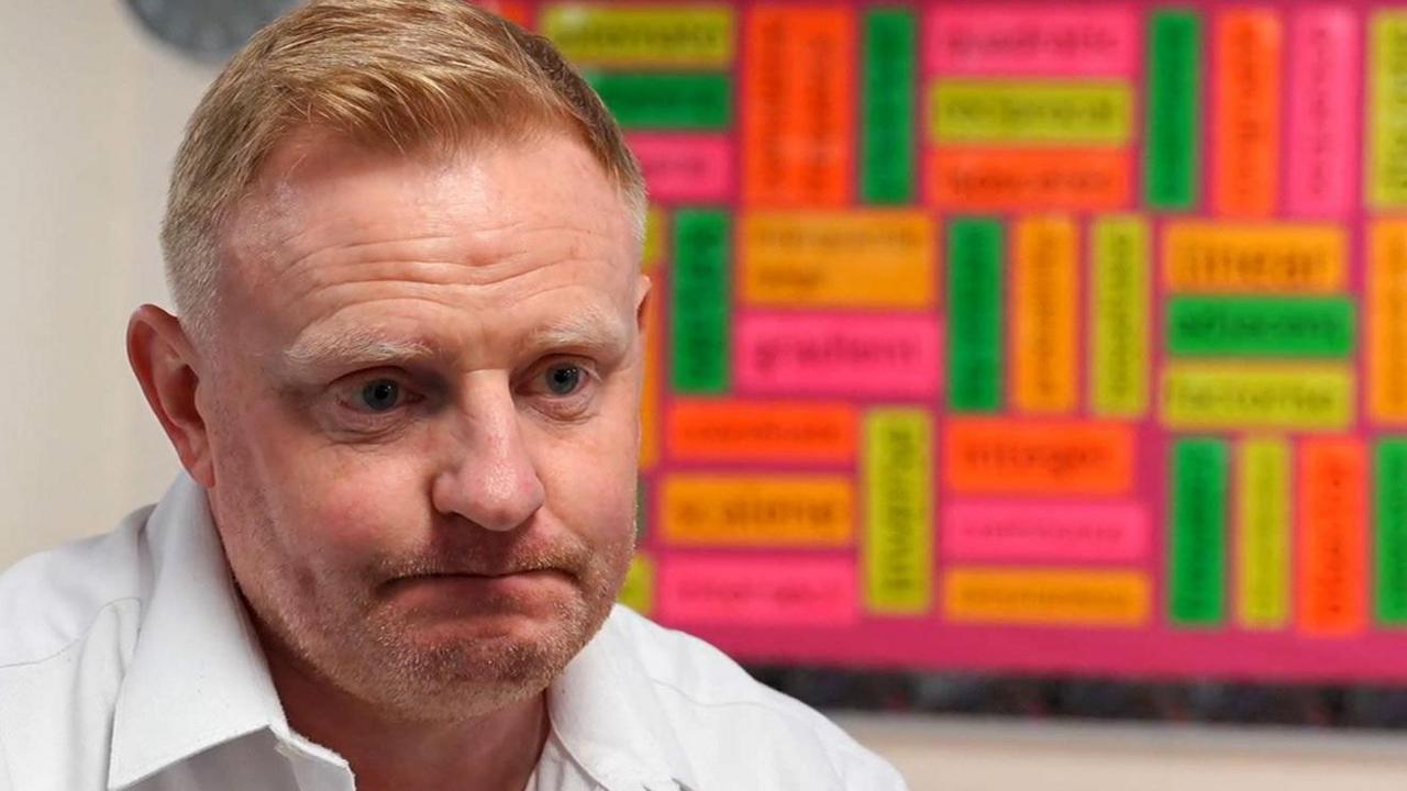 Paul Clark has short ginger-blond hair. He is looking slightly off-camera and wearing a white open-necked shirt. Behind him is a board with various rectangular luminous pieces of paper arranged vertically and horizontally. Each has words written on but they are blurred and not legible.