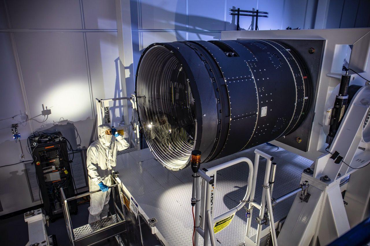 A person in a white cleanroom suit stands on a platform next to a large, cylindrical telescope with a wide, reflective lens. It is inside a large room with dim lighting, equipment, and safety railings.