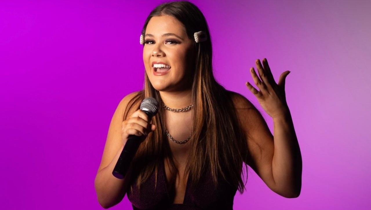 Isabella Giorgio-Jones with long dark hair holding a microphone in her right hand and putting her left hand aloft. She is in front of a purple background.