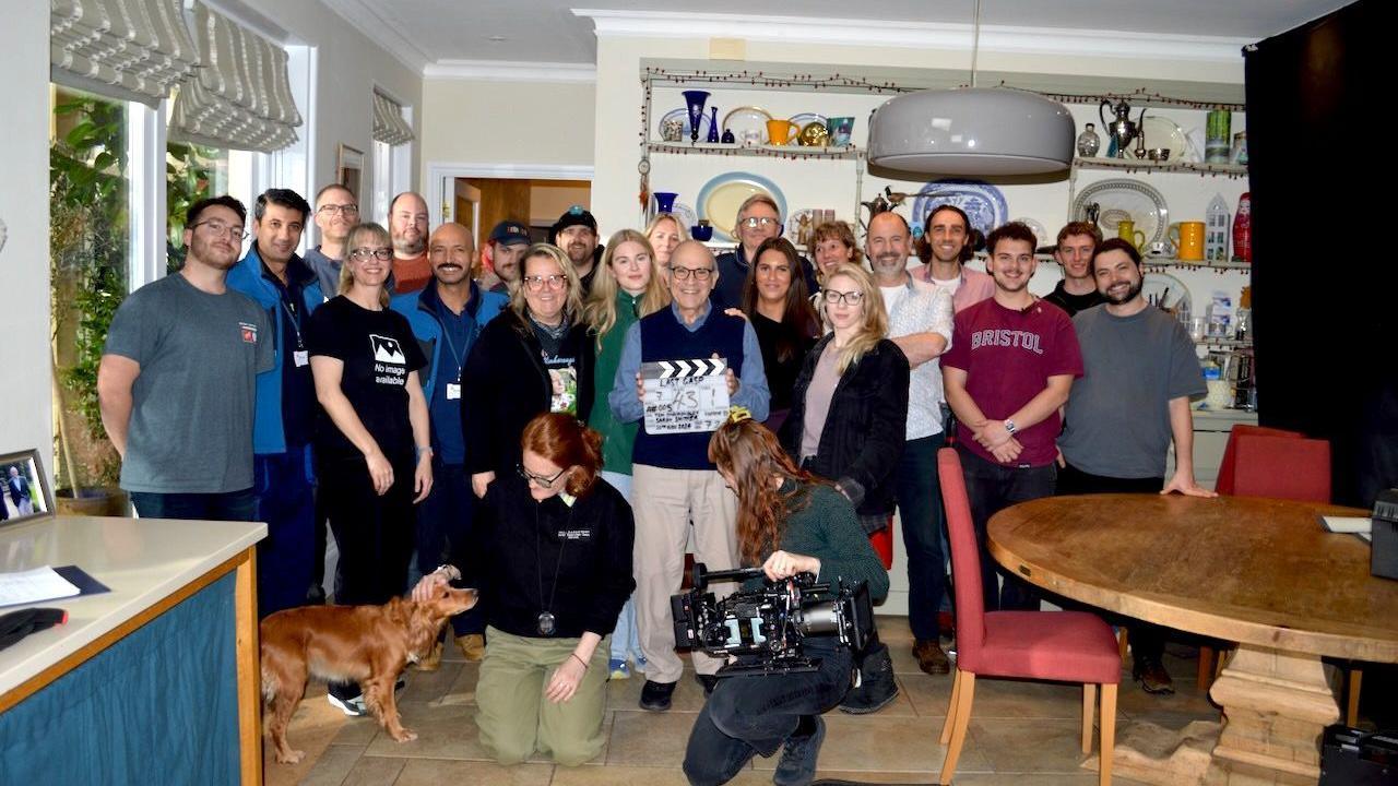 About 20 people pose together at a kitchen-diner film set, smiling at the camera. One man in the middle, Sir David Suchet, holds a clapperboard and a woman kneeling in front holds a large camera.