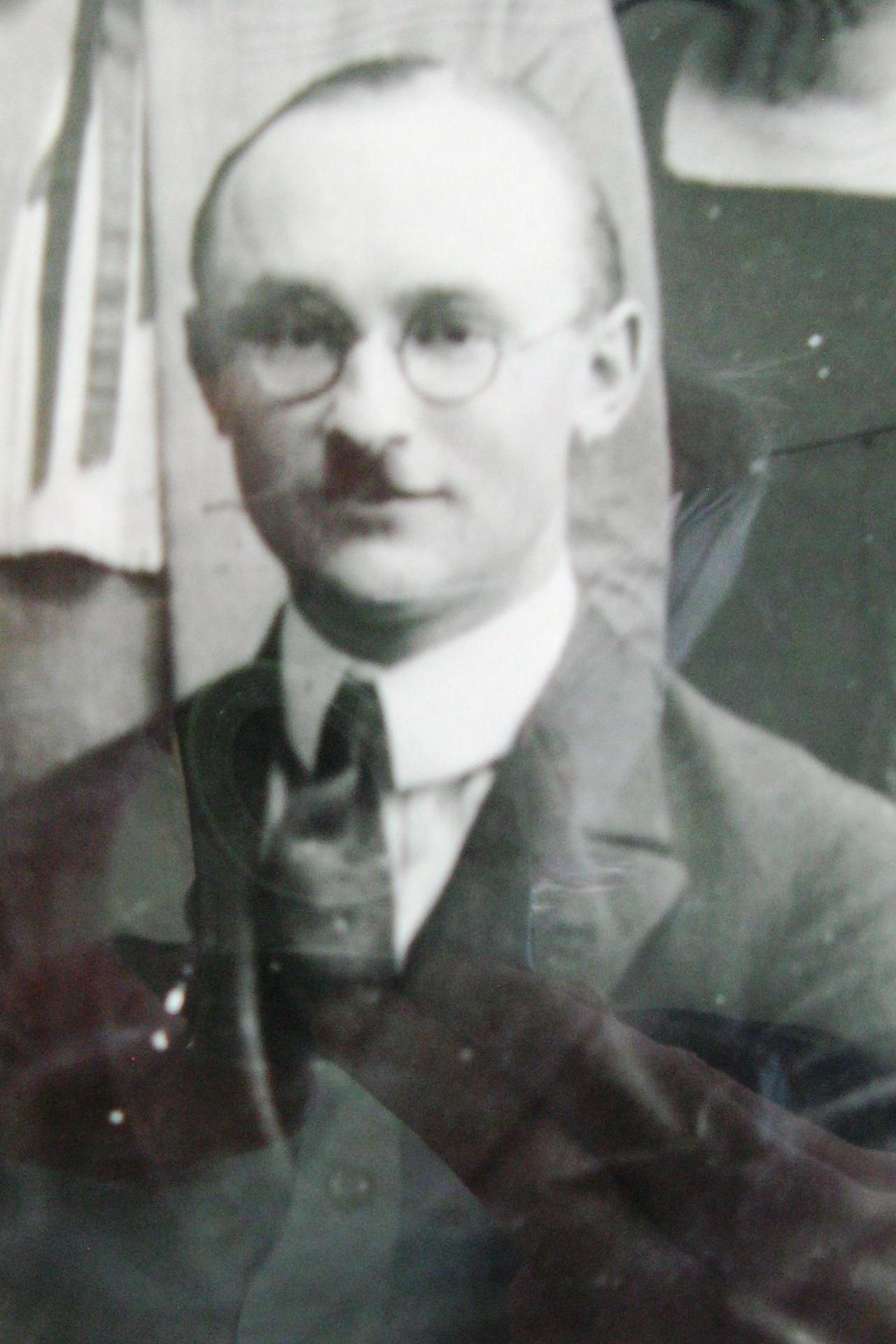 A seated Reinhard Haferkorn is shown in the foreground of a group photo. Wearing a suit and tie, he has rounded glasses and a Hitler-style moustache. It is a black and white image.