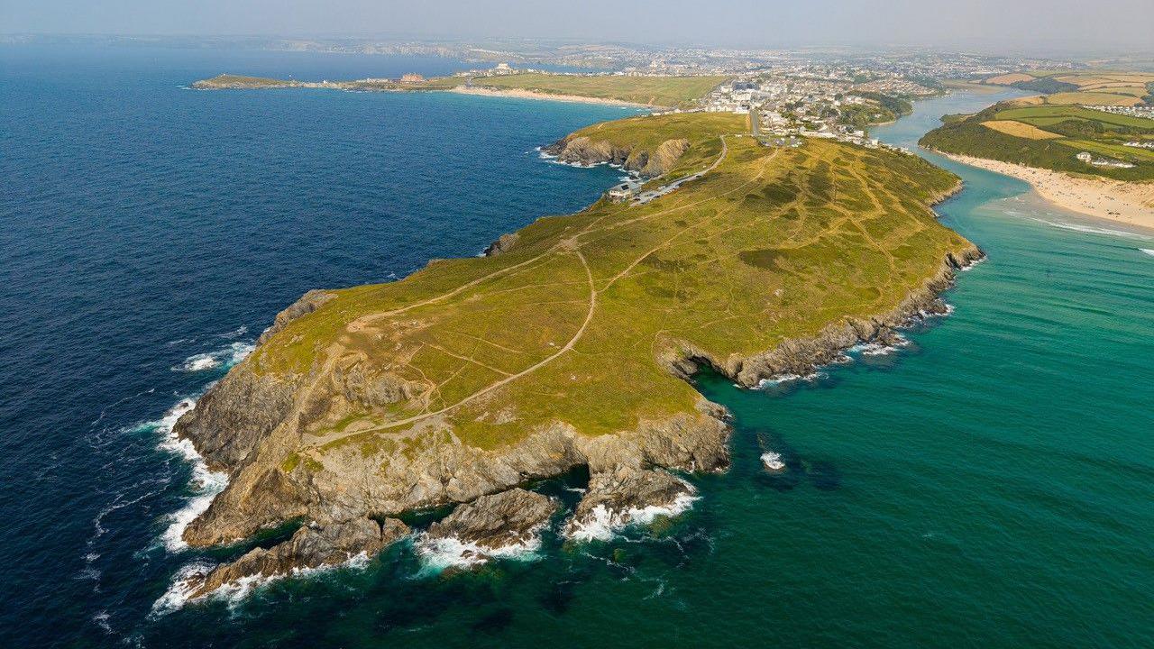 A drone shot above the sea showing the point of East Pentire Headland looking back to the rest of Newquay on a sunny day. The car park is barely visible as the drone is high in the sky. The headland is covered in scrub and grass and has footpaths crossing it. The sea to the left of the peninsula is blue and it is more green to the right. There is Crantock beach on West Pentire to the right behind which are fields and Fistral beach is visible to the left of East Pentire.