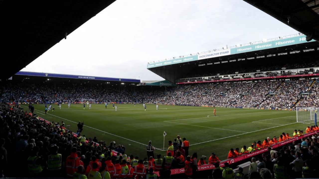 A photo of Leeds United's Elland Road stadium taken from inside the corner of the stadium. Fans are sat in the stand and players are on the pitch. The sky is cloudy.